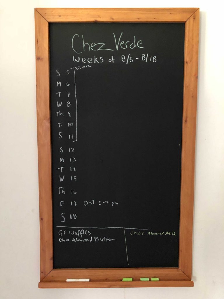 the chalkboard that Chris built. Chez Verde is written across the top. Below that is written weeks of 8/5 - 8/18. Along the left side is a list with a letter for the day of the week and a number for the date. Some dates have notations of events. Below the list of dates, two boxes drawn in chalk contain recipes to be made and groceries needed.