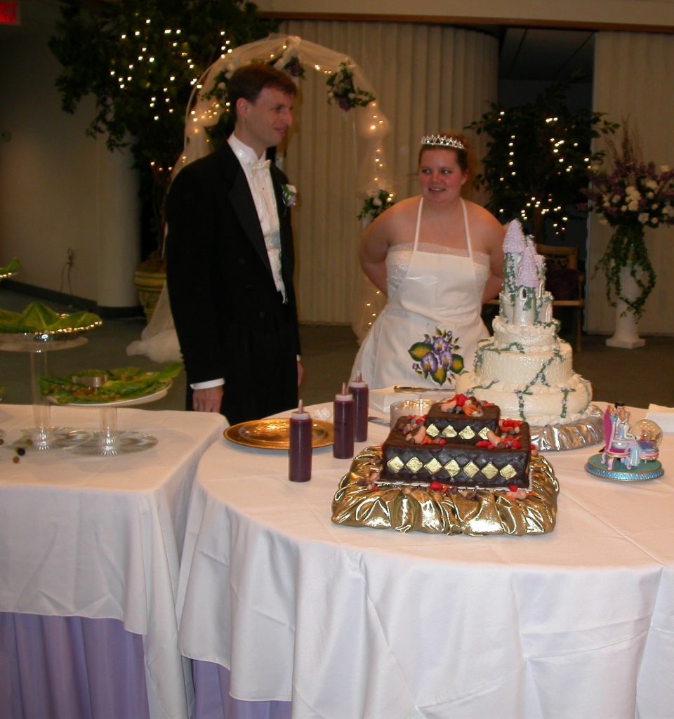 Chris and Karen standing behind the table holding their bride and groom cakes. Karen is tying an apron around her waist.